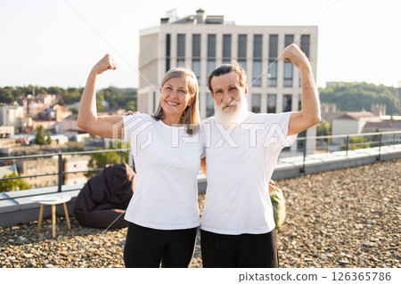 Elderly man and woman standing together on rooftop flexing arms, promoting active lifestyle and health awareness, showcasing positive emotions, cheerfulness, and physical fitness. 126365786