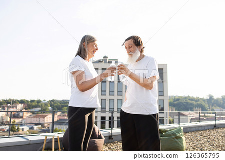 Elderly man and woman standing outside holding bottled water on rooftop after workout. Scene conveys feelings of health, companionship, and relaxation, with outdoor urban setting. 126365795