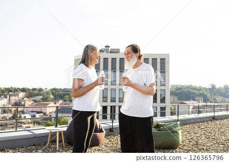 Senior man and woman in casual white shirts enjoying post-training conversation holding water outdoors. Setting urban rooftop location under sunlight creating cheerful and healthy atmosphere. 126365796