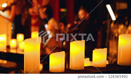Close-up of a row of lit candles in the foreground with a blurred background of a musical performance. Chamber music concert by candlelight. Close-up of a row of lit candles in the foreground with a blurred background of a musical performance. Chamber music concert by candlelight. 126365992