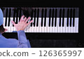 Close-up view of pianist's hand stretched across black and white piano keys, wearing light blue sleeve, partial view of blue beanie visible, shot from above against dark background. 126365997
