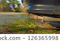 Close-up of a car's exhaust pipe emitting smoke. The car is parked on a grassy area with a sidewalk and trees in the background. Environmental Issues. Greenhouse gases. 126365998