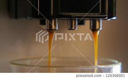 A close-up of an espresso machine with two spouts pouring espresso into a glass cup. The espresso is a dark brown color and is dripping into the cup. 126366001