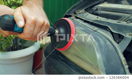A close-up a person using a power drill with a buffing attachment to restore a cloudy and discolored car headlight. The headlight lens is being polished to improve its clarity and appearance. A close-up a person using a power drill with a buffing attachment to restore a cloudy and discolored car headlight. The headlight lens is being polished to improve its clarity and appearance. 126366004