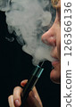 Side profile of a woman's face exhaling a cloud of vapor from a vape device clutched between her fingers. Swirling patterns of vapor on a black background with dramatic lighting. 126366136