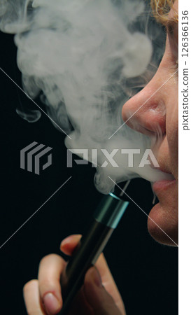Side profile of a woman's face exhaling a cloud of vapor from a vape device clutched between her fingers. Swirling patterns of vapor on a black background with dramatic lighting. 126366136