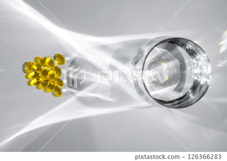 Yellow vitamin D or fish oil capsules are scattered near a glass of water, casting intriguing shadows on a white surface, creating an aesthetically pleasing composition 126366283