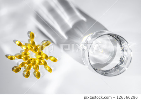 vitamin D or Fish oil yellow transparent capsules forming a sun shape alongside a glass of water create playful shadows on a white background, emphasizing themes of health, wellness, and nutrition 126366286
