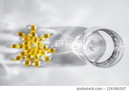 vitamin D or Fish oil yellow transparent capsules forming a sun shape alongside a glass of water create playful shadows on a white background, emphasizing themes of health, wellness, and nutrition 126366287