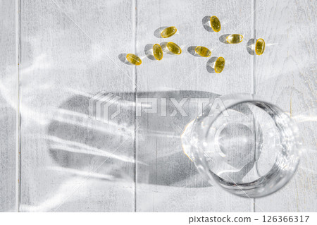 Yellow transparent capsules with vitamins or supplements and a glass of water are lying on a white wooden table, illuminated by a ray of sunlight casting shadows Yellow transparent capsules with vitamins or supplements and a glass of water are lying on a white wooden table, illuminated by a ray of sunlight casting shadows 126366317