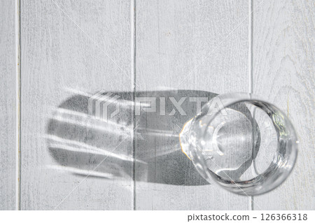 Glass of water casting a long shadow on a white wooden table, illuminated by natural light, forming an abstract and minimalist composition that emphasizes simplicity and clarity 126366318