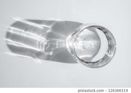 Glass of water resting on a white surface, creating an elongated and distorted shadow through the refraction of light, highlighting transparency and purity in a minimalistic setting 126366319