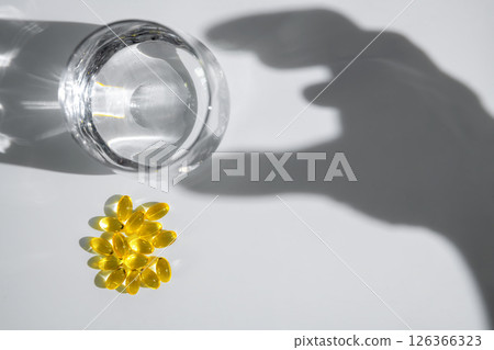 Yellow transparent capsules or pills or vitamins or supplements are near glass of water with hand shadow reaching for them on white background, representing medicine and healthcare Yellow transparent capsules or pills or vitamins or supplements are near glass of water with hand shadow reaching for them on white background, representing medicine and healthcare 126366323
