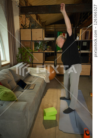 Elderly man standing on yoga mat with one arm raised overhead, performing exercise in living room. Laptop, yoga blocks visible. Elderly man standing on yoga mat with one arm raised overhead, performing exercise in living room. Laptop, yoga blocks visible. 126366327