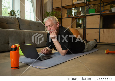 Elderly man lying on yoga mat and looking at laptop, with his chin resting on his hand. A water bottle and dumbbell on floor nearby. Elderly man lying on yoga mat and looking at laptop, with his chin resting on his hand. A water bottle and dumbbell on floor nearby. 126366328