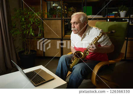 Elderly man with mustache sitting in green armchair, holding saxophone and looking at laptop, online music lesson 126366330