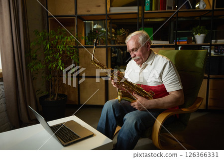 Elderly man with mustache sitting in green armchair, holding saxophone and looking at laptop, online music lesson. 126366331