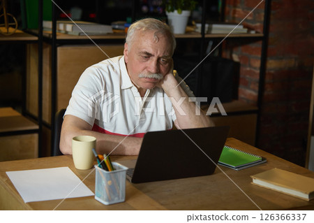 Senior man working on laptop at wooden desk in dark office. Concentrated face, deeply involved in learning process with modern technologies. 126366337