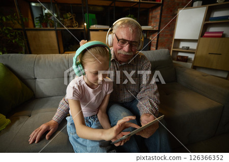 Focused child girl and elderly man sitting on couch in headphones and interacting with digital tablet. Modern learning and gadgets Focused child girl and elderly man sitting on couch in headphones and interacting with digital tablet. Modern learning and gadgets 126366352