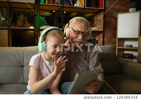 Smiling child girl and senior man, bot in headphones, sitting on couch and waving towards tablet, having online video call in cozy living room. 126366358