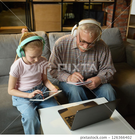 Elderly man and girl wearing headphones, using tablets and writing notes during online learning in cozy living room. Elderly man and girl wearing headphones, using tablets and writing notes during online learning in cozy living room. 126366402