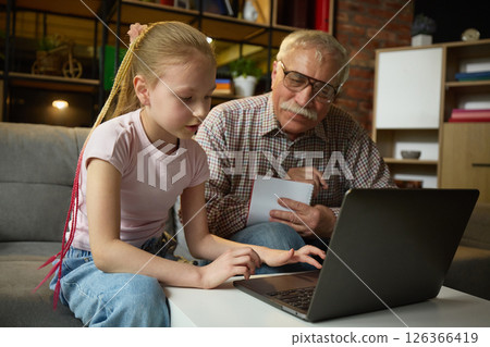 Child girl focusing on laptop while elderly man observing with attention and taking notes during home learning session. 126366419