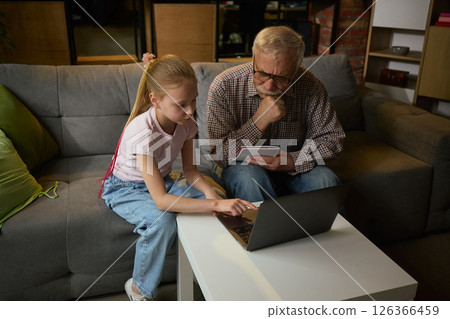 Young girl helping elderly man with laptop while studying together on sofa in cozy home setting. Two generations, support and development. 126366459