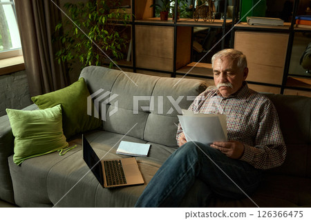 Senior man lying back on sofa and reading papers next to laptop and notebook in cozy living room. Comfortable education for seniors. 126366475