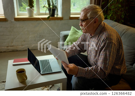 Focused elderly man working on laptop at home, holding papers, sitting on sofa in living room with coffee cup on table. 126366482