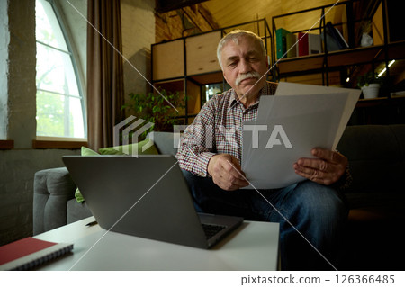 Focused elderly man with mustache sitting on sofa in modern living room, looking at papers, with laptop on table 126366485
