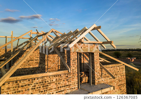 Private residential house with wooden roof frame structure under construction. Unfinished brick building under development. Private residential house with wooden roof frame structure under construction. Unfinished brick building under development. 126367085