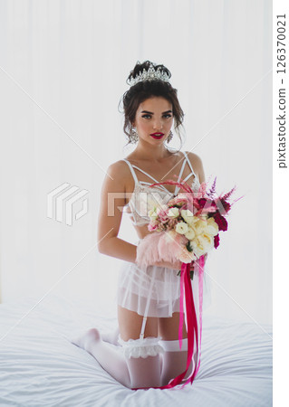 Seductive slender young bride in a sexy lingerie and white stockings posing on a bed with her bridal bouquet 126370021