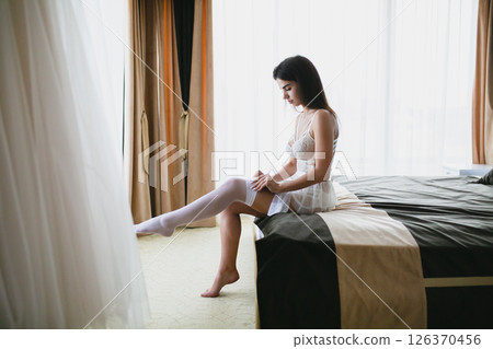 Seductive slim bride in sexy lingerie on bed in hotel room puts on white stockings 126370456