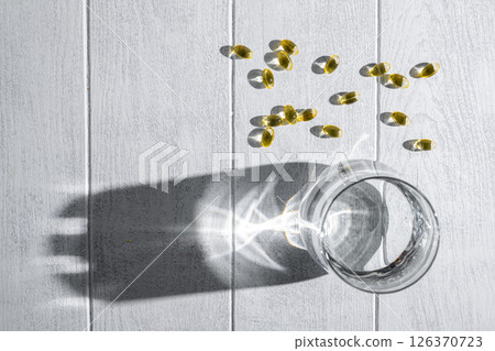 Yellow transparent capsules with vitamins or supplements and a glass of water are lying on a white wooden table, illuminated by a ray of sunlight casting shadows Yellow transparent capsules with vitamins or supplements and a glass of water are lying on a white wooden table, illuminated by a ray of sunlight casting shadows 126370723