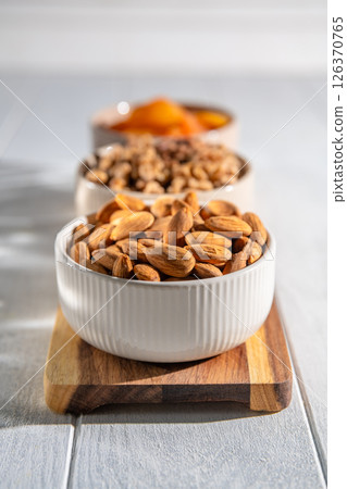 Three bowls of healthy snacks including dried apricots, walnuts, and almonds are arranged on a wooden board, offering a nutritious and delicious treat 126370765