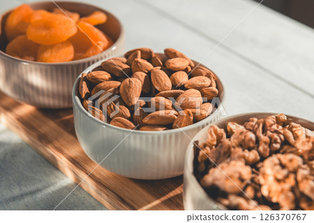 Three bowls of healthy snacks including dried apricots, walnuts, and almonds are arranged on a wooden board, offering a nutritious and delicious treat 126370767