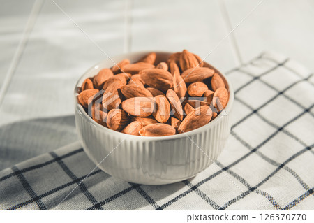 Shelled almonds are piled high in a small white bowl resting on a checkered cloth, creating a simple yet appealing image of healthy snacking Shelled almonds are piled high in a small white bowl resting on a checkered cloth, creating a simple yet appealing image of healthy snacking 126370770