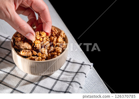 Woman's hand reaches into a bowl of walnuts, picking one from the pile on a checkered cloth, ready for a healthy snack or ingredient in a recipe Woman's hand reaches into a bowl of walnuts, picking one from the pile on a checkered cloth, ready for a healthy snack or ingredient in a recipe 126370785
