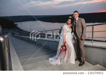 Young beautiful couple of newlywed posing in the evening against the background of an atmospheric panorama and stunning sunset Young beautiful couple of newlywed posing in the evening against the background of an atmospheric panorama and stunning sunset 126370952