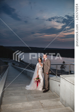 Young beautiful couple of newlywed posing in the evening against the background of an atmospheric panorama and stunning sunset Young beautiful couple of newlywed posing in the evening against the background of an atmospheric panorama and stunning sunset 126370954