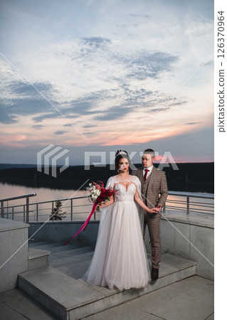 Young beautiful couple of newlywed posing in the evening against the background of an atmospheric panorama and stunning sunset Young beautiful couple of newlywed posing in the evening against the background of an atmospheric panorama and stunning sunset 126370964