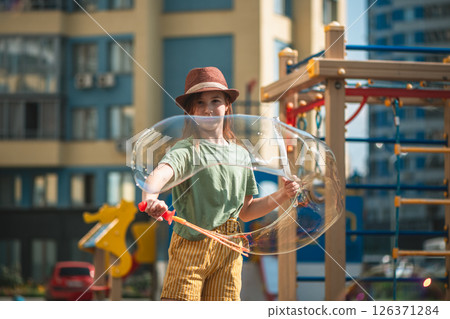 Cute school-age girl blows big soap bubbles in the courtyard of a multi-storey building 126371284