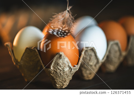 Brown and white chicken eggs are nestled in a cardboard carton, a speckled feather resting gently atop one, creating a rustic, farm-fresh still life 126371455