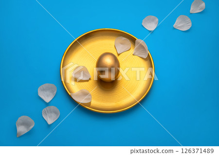 Luxurious golden easter egg rests on a golden plate adorned with delicate white petals. Creating a sophisticated easter scene against a vibrant blue backdrop with water droplets. Viewed from above Luxurious golden easter egg rests on a golden plate adorned with delicate white petals. Creating a sophisticated easter scene against a vibrant blue backdrop with water droplets. Viewed from above 126371489