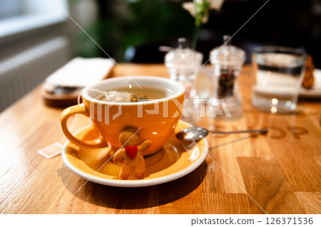 Gingerbread man with a red heart is hugging a yellow cup of tea, sitting on a saucer with a spoon, on a wooden cafe table, creating a cozy and festive atmosphere Gingerbread man with a red heart is hugging a yellow cup of tea, sitting on a saucer with a spoon, on a wooden cafe table, creating a cozy and festive atmosphere 126371536