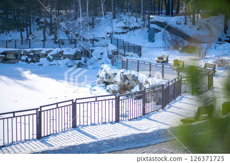 Winding metal fences encircle a frozen pond blanketed in snow, creating a serene winter park scene. Benches and trees enhance the tranquil atmosphere, inviting peaceful moments 126371725