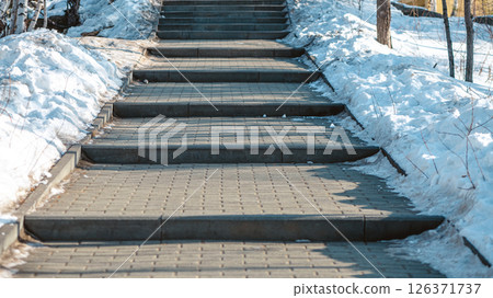 Concrete block paved path bordered by a metal fence and snow going slightly uphill towards a stairway in a park with pine and birch trees during winter Concrete block paved path bordered by a metal fence and snow going slightly uphill towards a stairway in a park with pine and birch trees during winter 126371737