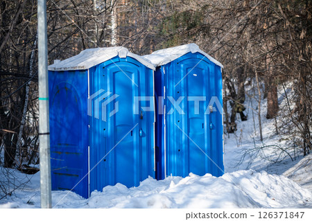 Two blue portable toilet cabins are standing on a snowy ground in winter, surrounded by leafless trees and bushes, providing necessary facilities in outdoor environments 126371847