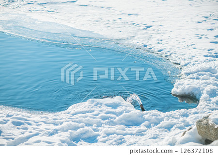 Water gushing from a pipe into a vibrant blue polynya amidst a snow-covered, icy landscape, creating a striking contrast between the cold environment and flowing water Water gushing from a pipe into a vibrant blue polynya amidst a snow-covered, icy landscape, creating a striking contrast between the cold environment and flowing water 126372108