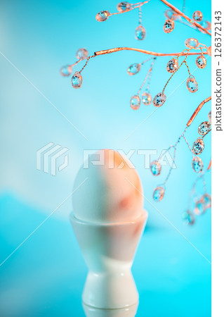 Easter egg in a white egg cup with the shadow of a decorative branch projected on it, creating a beautiful and festive atmosphere on a gradient blue background 126372143
